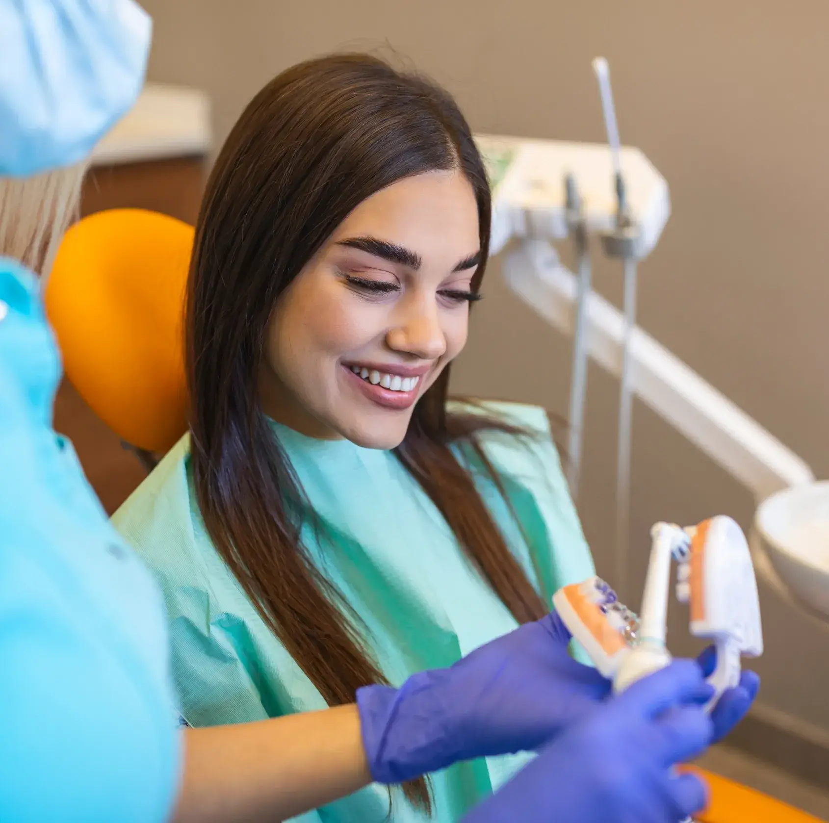 Smiling woman in dental chair wearing a green bib looking at a dental model held by a gloved dentist.