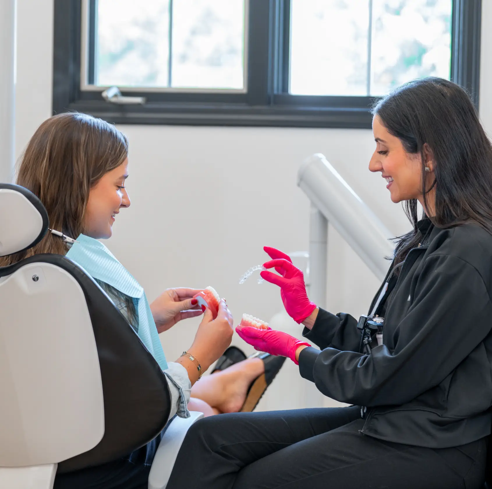 Smiling woman in dental chair wearing a green bib looking at a dental model held by a gloved dentist.