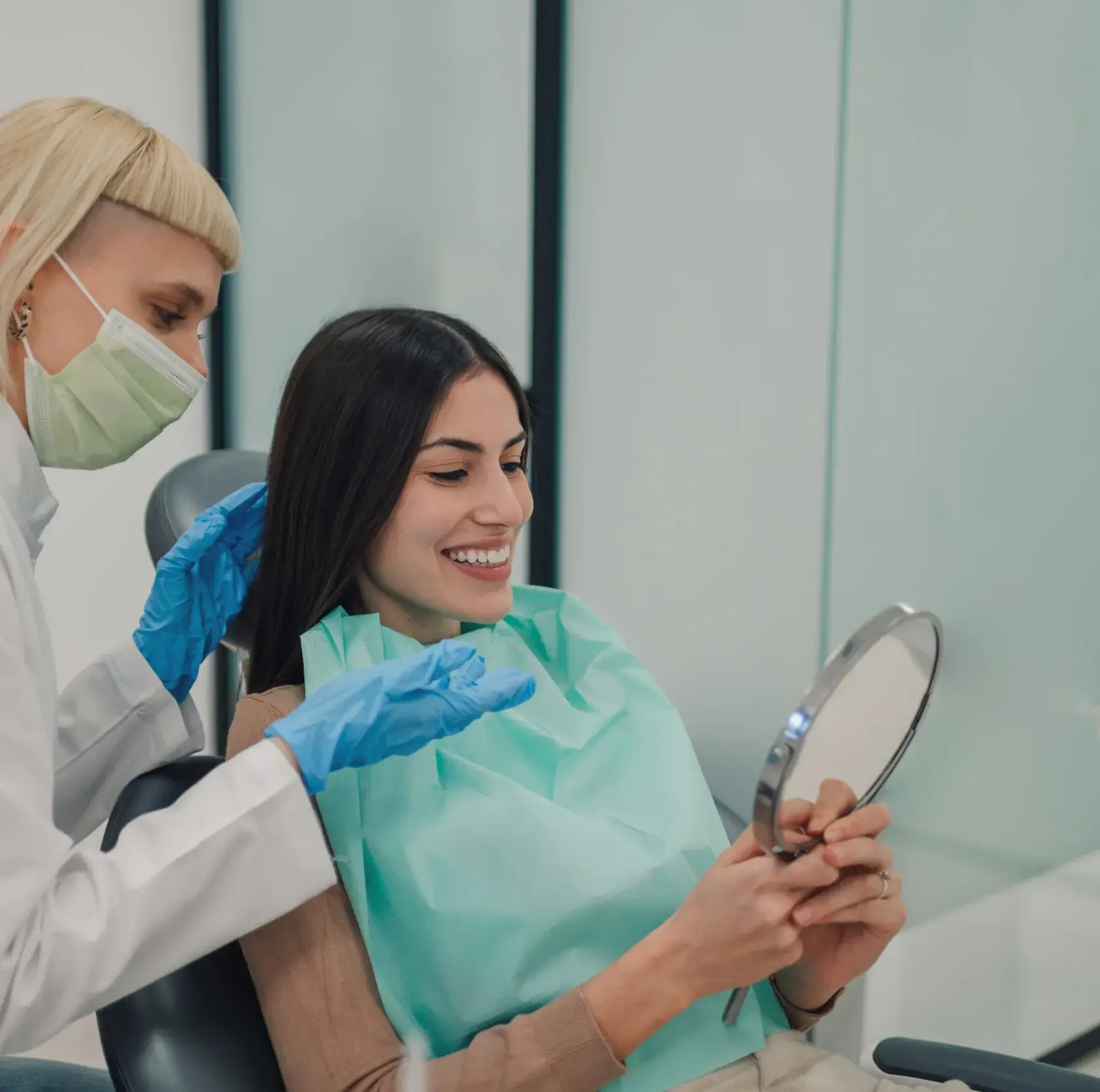 Dentist wearing mask and gloves showing a smiling patient her teeth in a handheld mirror.