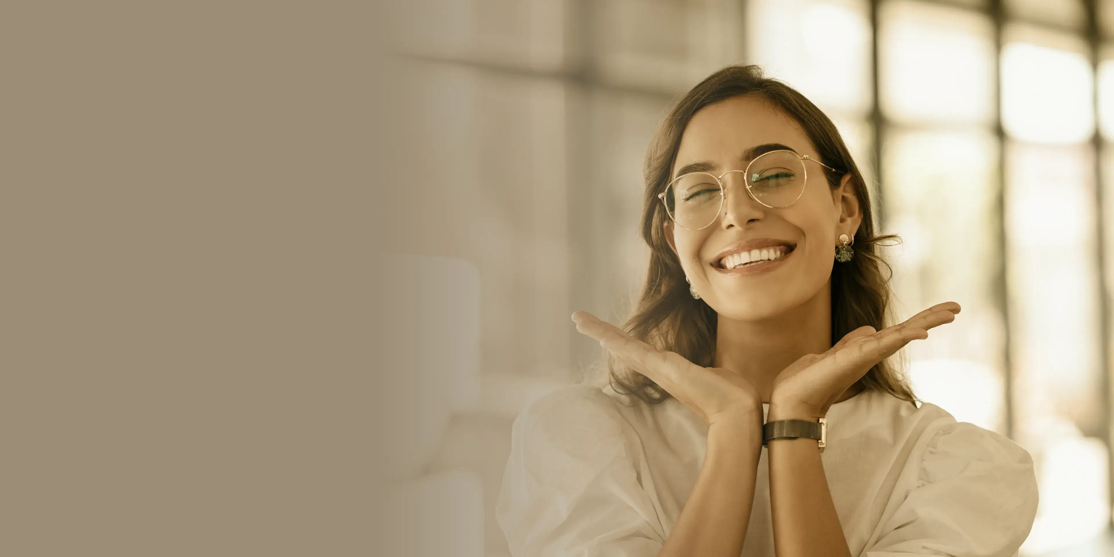 Smiling woman with glasses and earrings posing with hands under chin in bright indoor setting.
