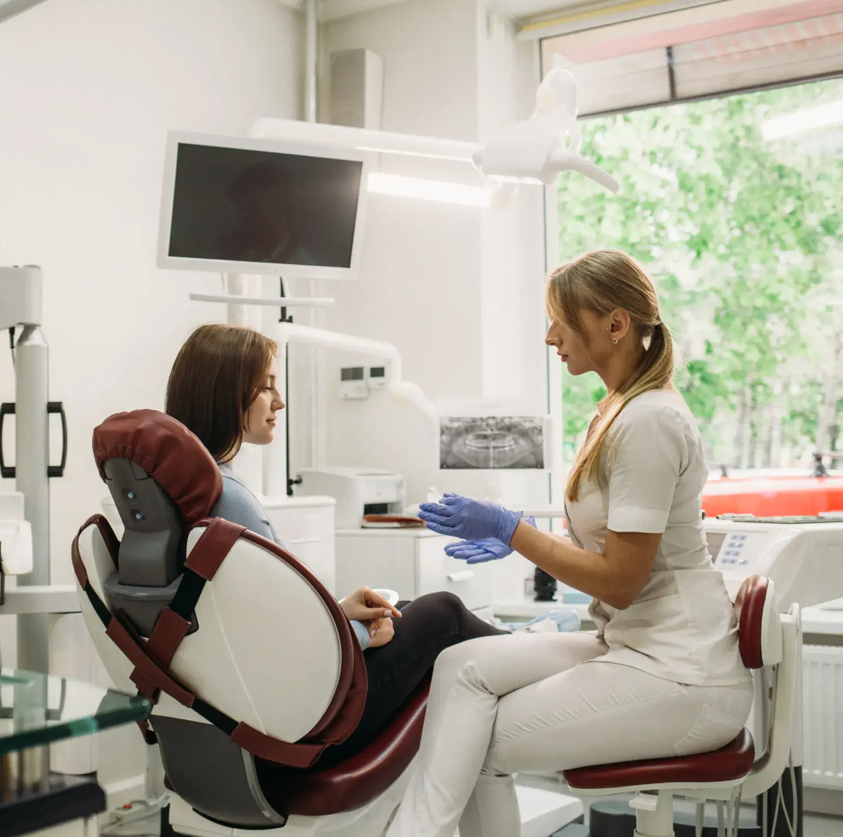 Dentist in white uniform wearing blue gloves explaining clear aligners to a woman sitting in a dental chair in a bright clinic.