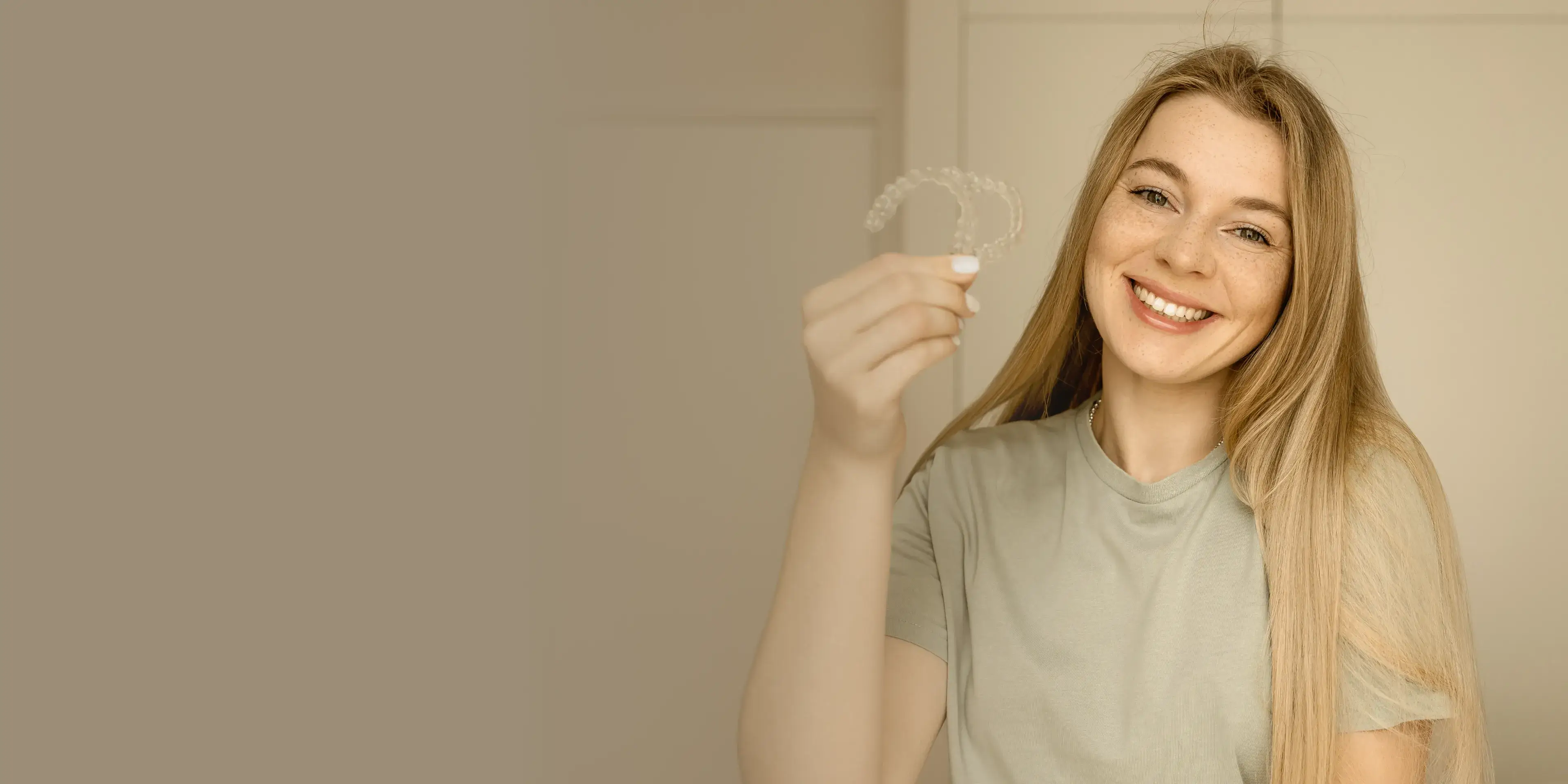 Smiling young woman with long blonde hair holding clear dental aligners.