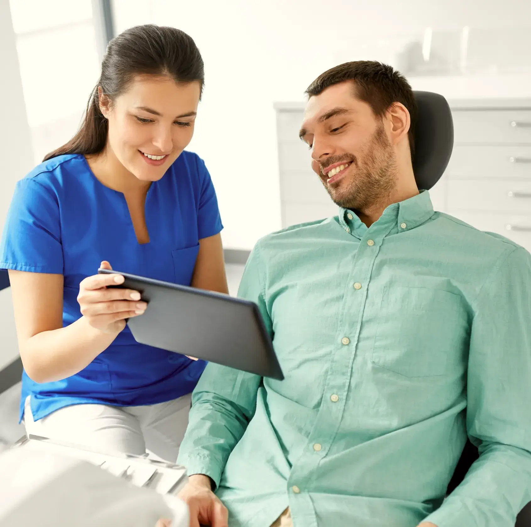 Smiling dental professional showing a tablet to a male patient seated in a dental chair.