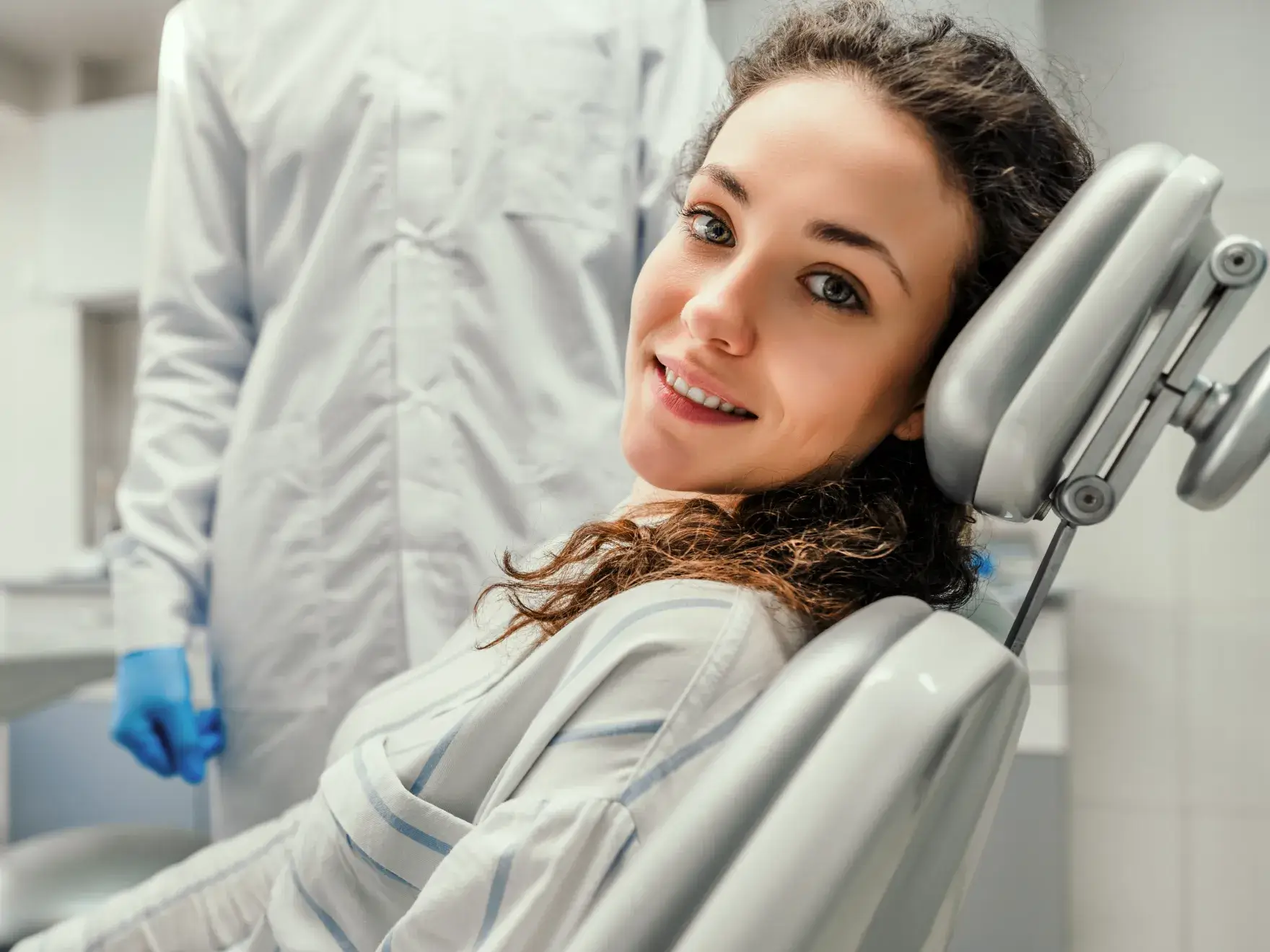 Smiling woman with curly hair reclining in a dental chair, with a person in a white coat and blue gloves standing behind her.