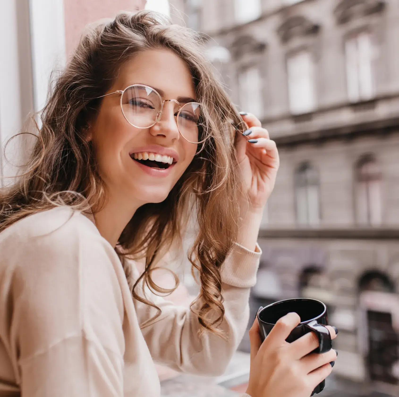 Smiling woman with glasses holding a black mug, sitting near a window with blurred city buildings in the background.