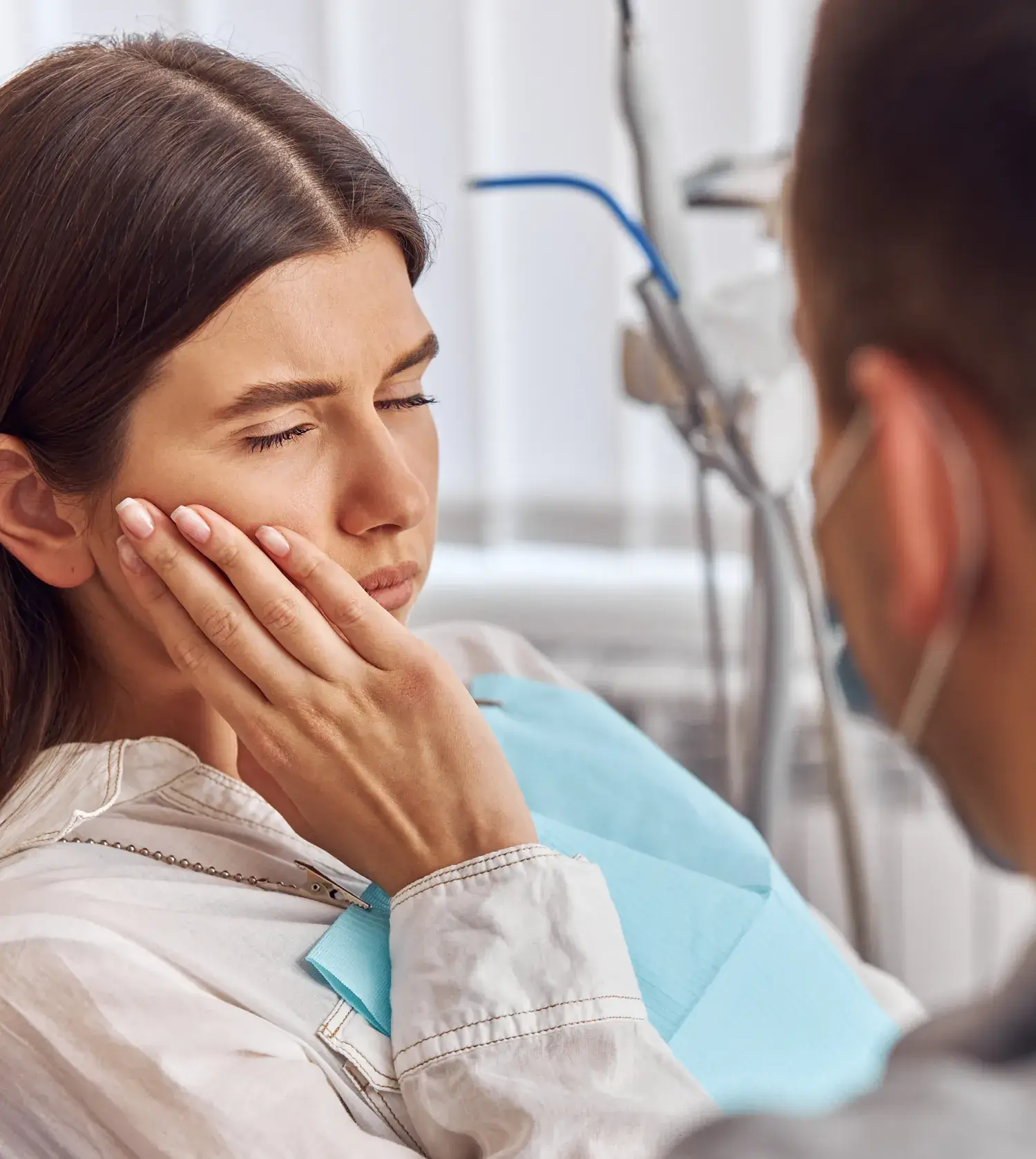 Woman in a dental chair holding her cheek in pain while a dentist wearing a mask attends to her.