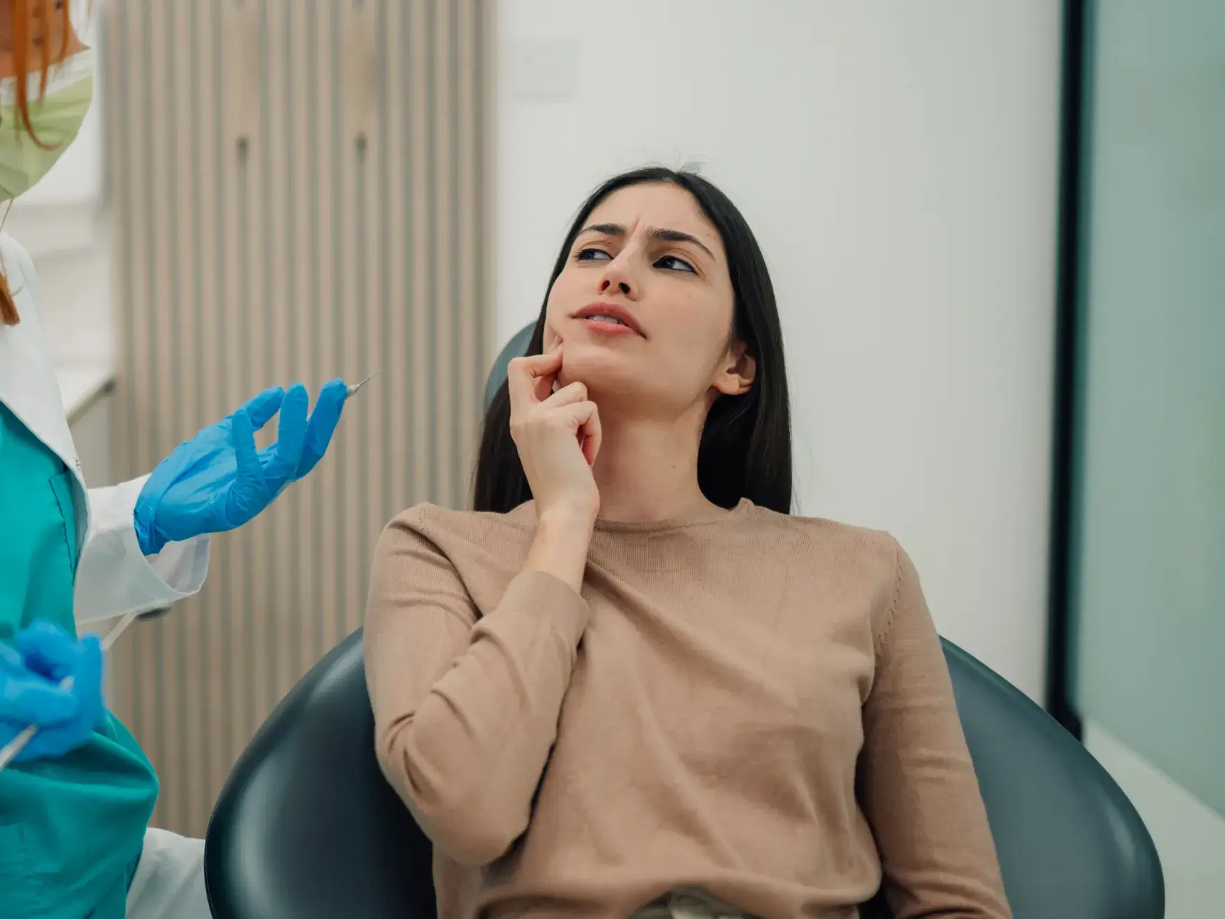 Woman sitting in a dental chair touching her cheek with a concerned expression while a dentist wearing blue gloves holds a dental tool.