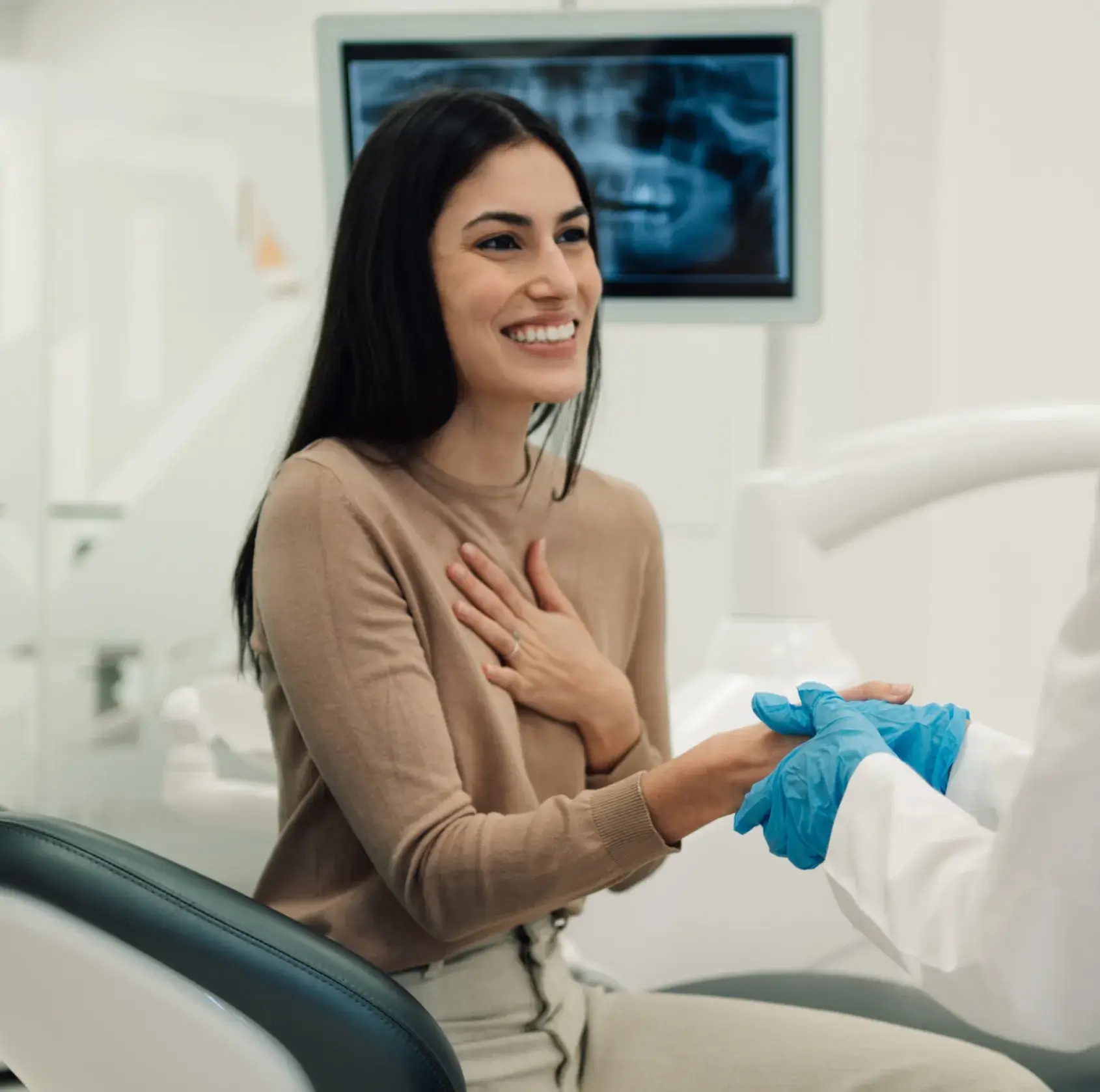 Smiling woman sitting in a dental chair holding hands with a dentist wearing blue gloves, with a dental X-ray displayed in the background.