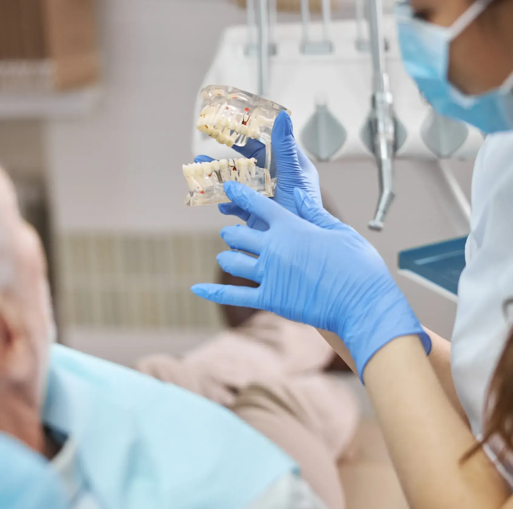 Dentist wearing blue gloves holding a dental model while consulting an elderly patient.