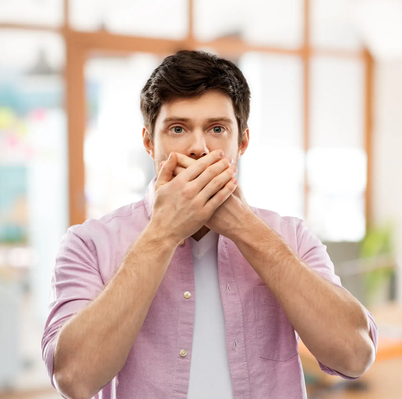 Young man wearing a pink shirt covering his mouth with both hands in a bright indoor setting.