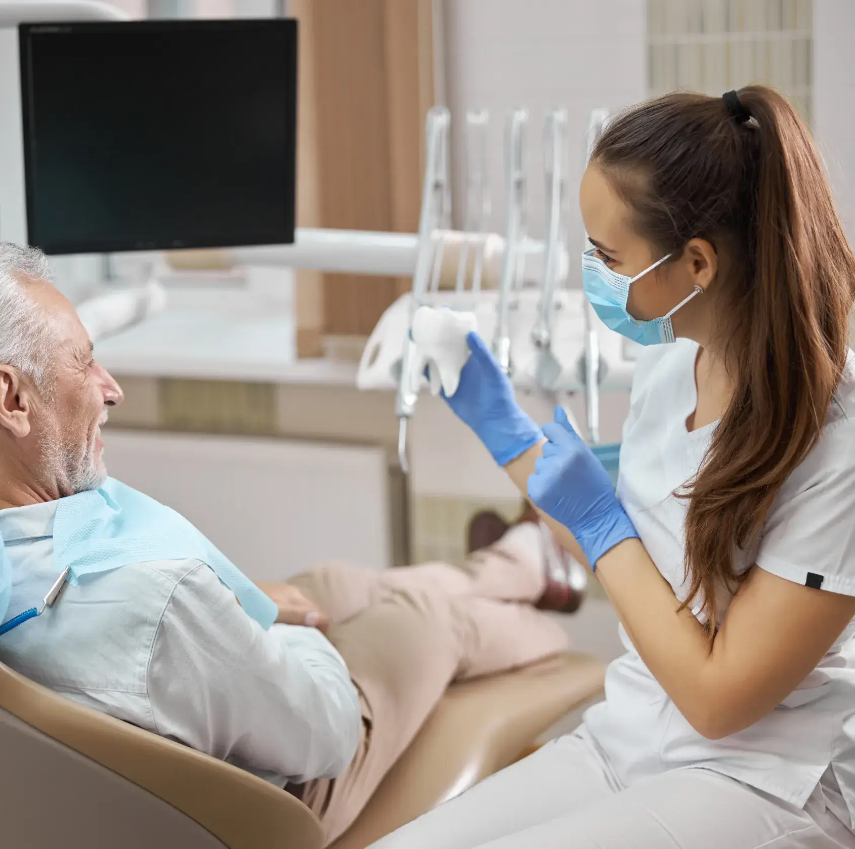 Dental professional wearing a mask and gloves explaining a tooth model to an elderly male patient reclining in a dental chair.