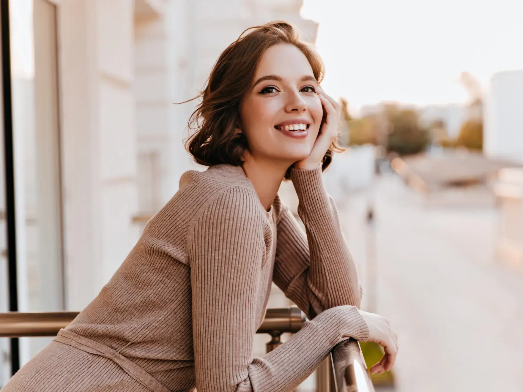 Smiling woman with short brown hair wearing a beige sweater leaning on a balcony railing outdoors.