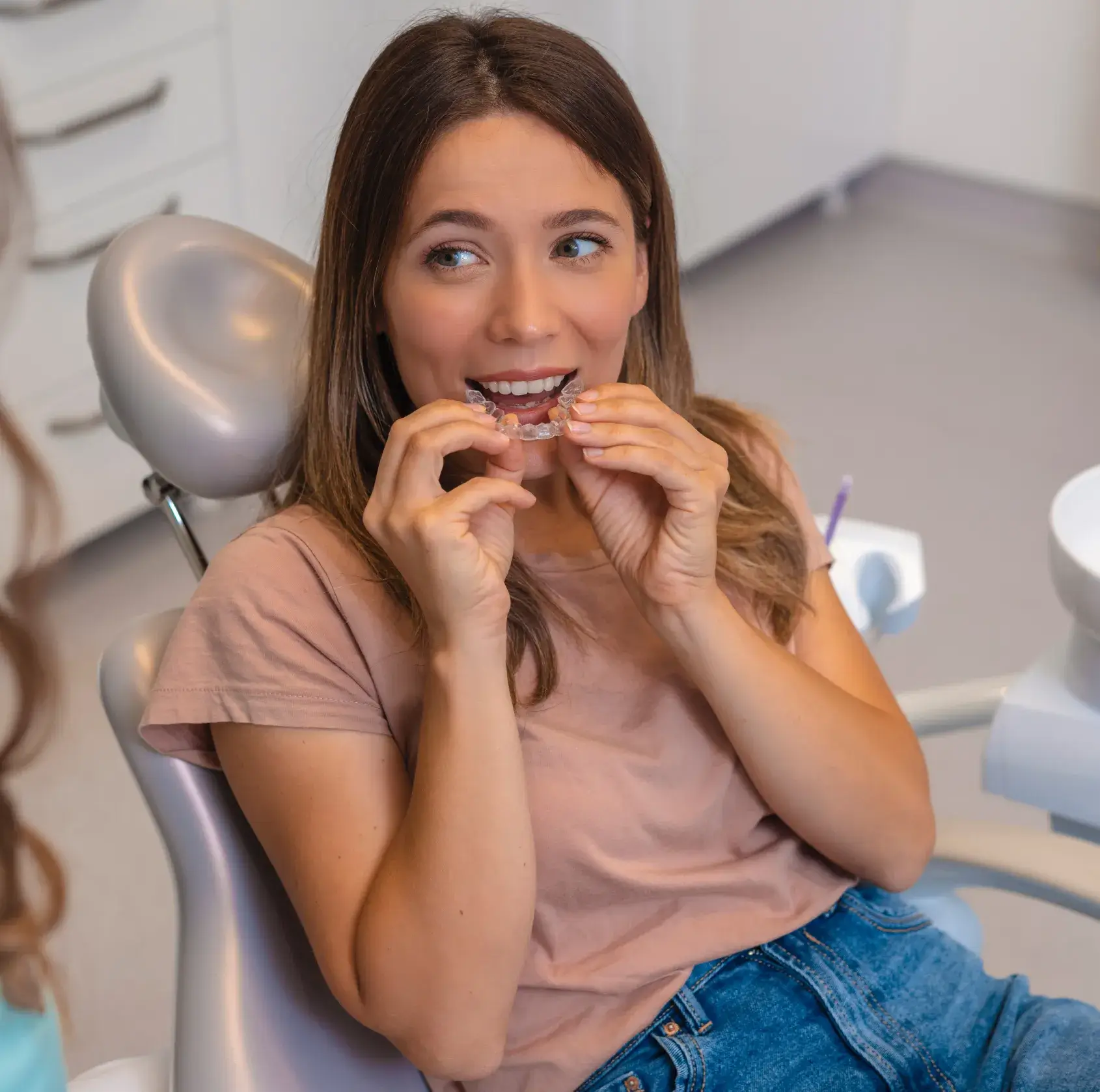 Woman sitting in a dental chair putting clear dental aligners in her mouth.