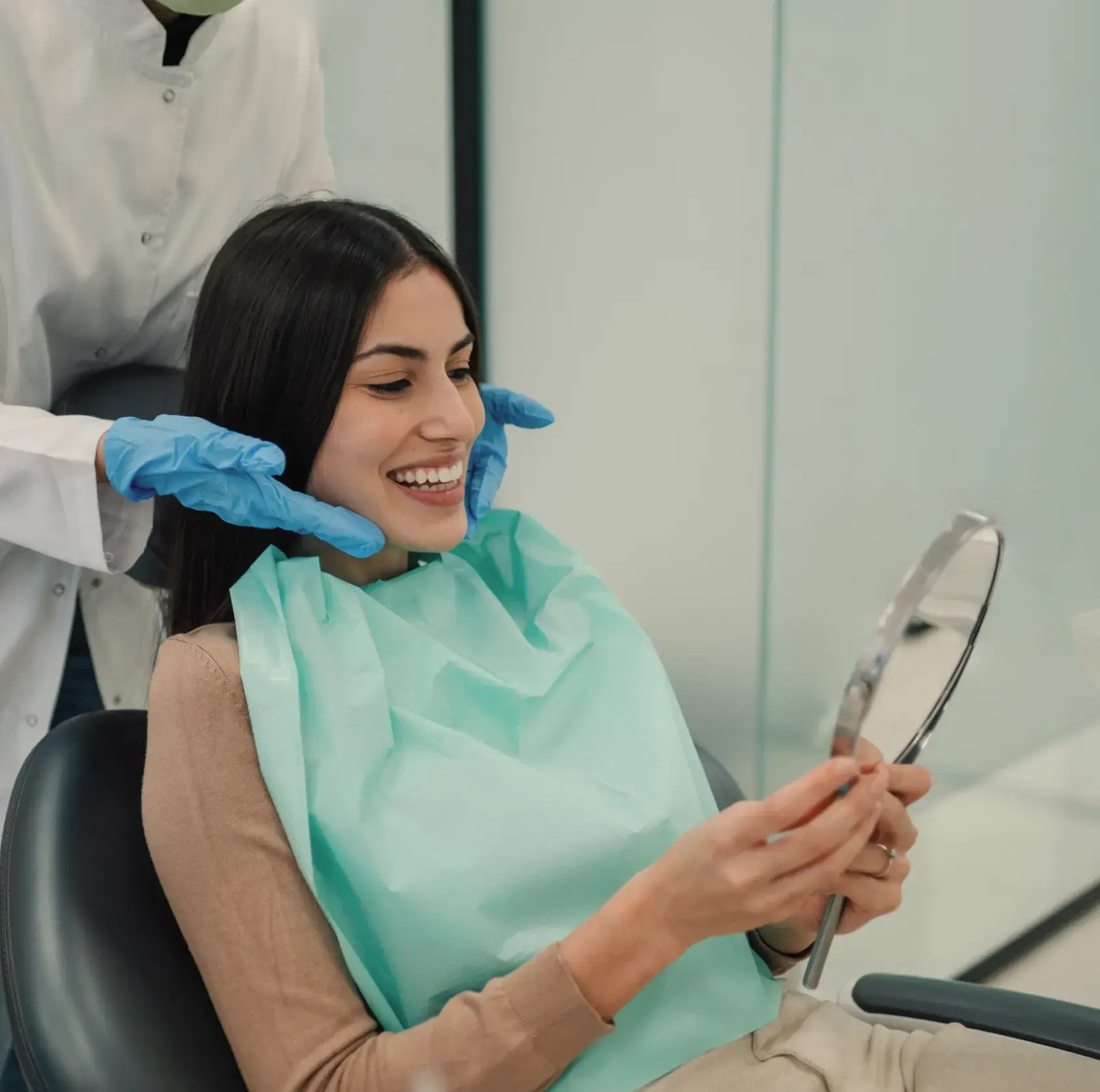 Smiling woman in dental bib holding a mirror while a professional wearing blue gloves examines her face.