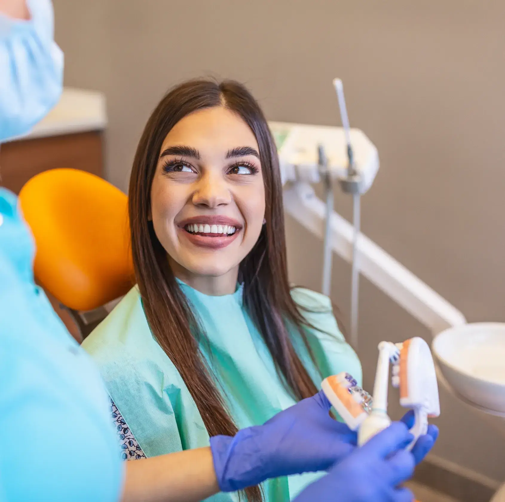Smiling woman in dental chair looking at a dentist demonstrating teeth brushing with dental models.