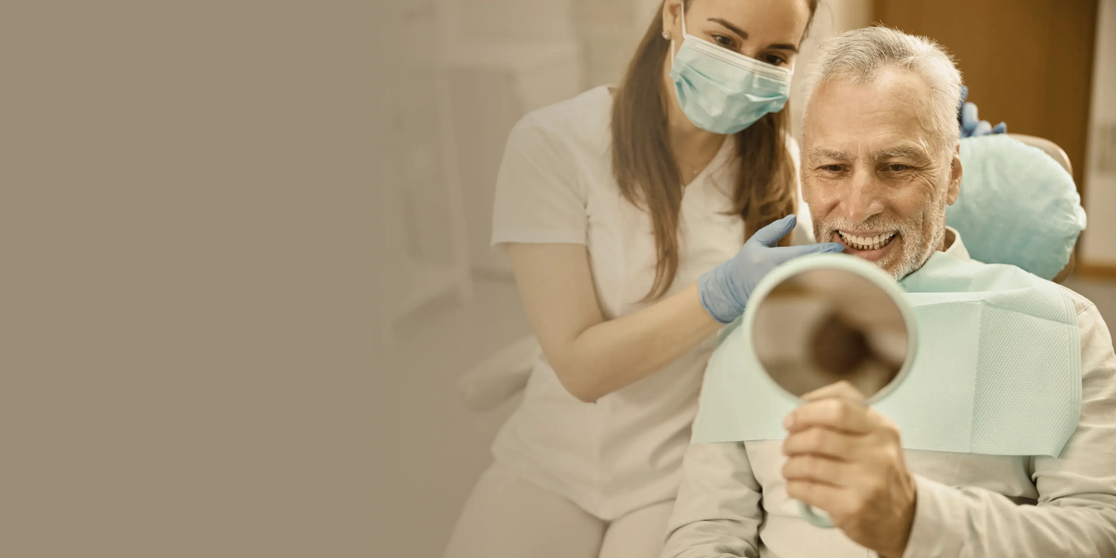 Elderly man sitting in dental chair smiling while looking at his teeth in a hand mirror, dentist with mask and gloves behind him.