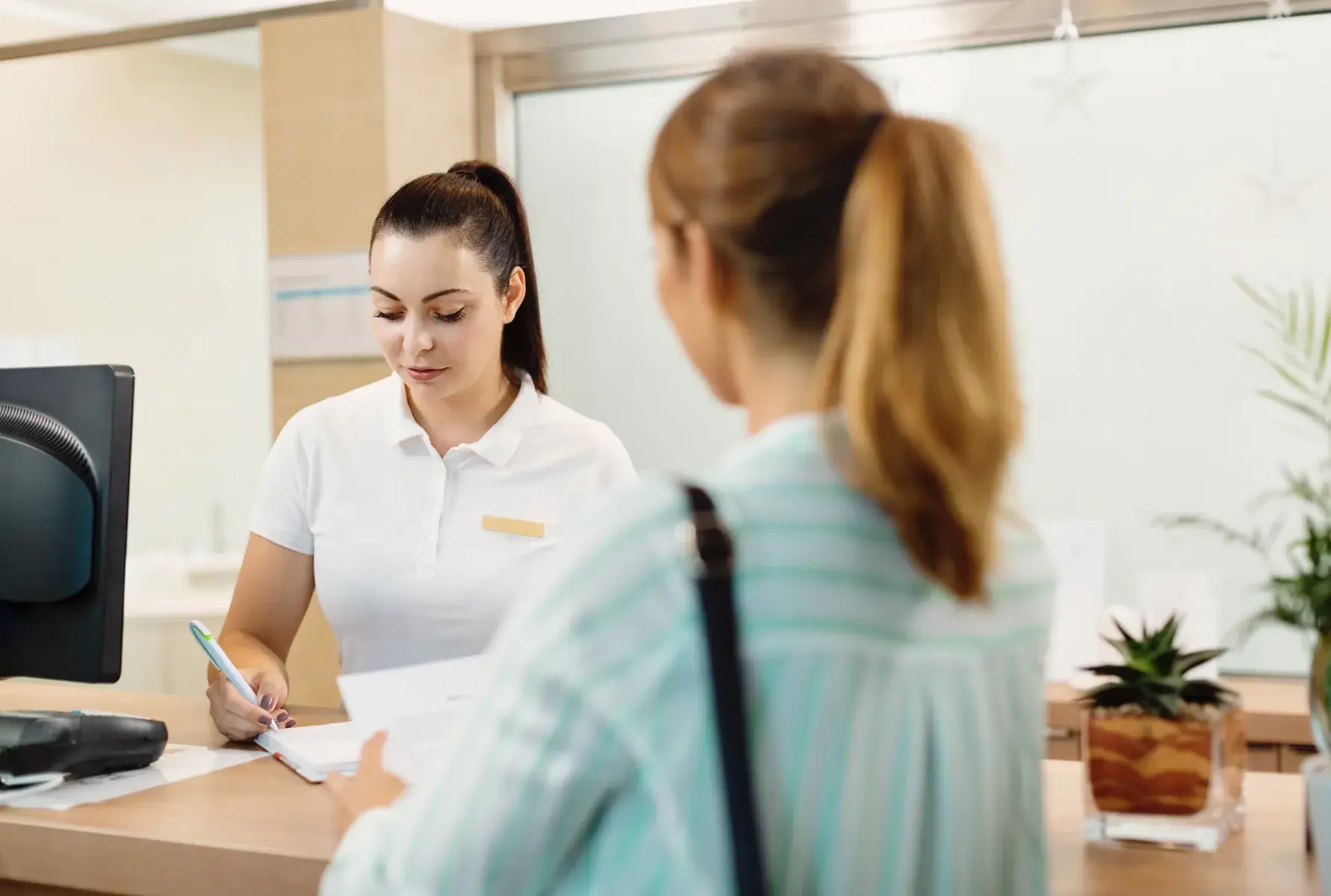 Receptionist in white shirt writing on a clipboard while a woman in a striped shirt waits at the counter.