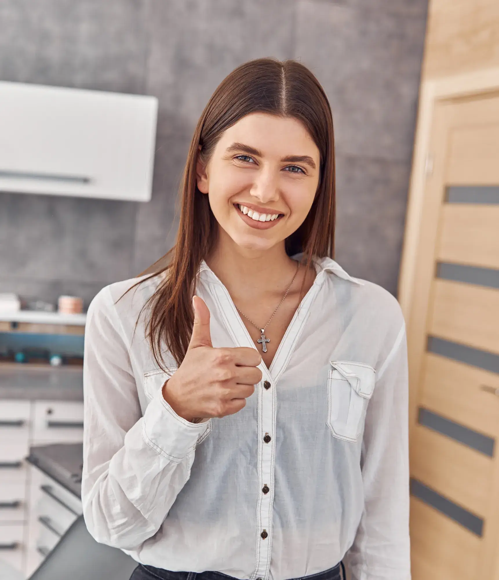 Smiling woman with long brown hair wearing a white button-up shirt showing a thumbs up in a modern kitchen.