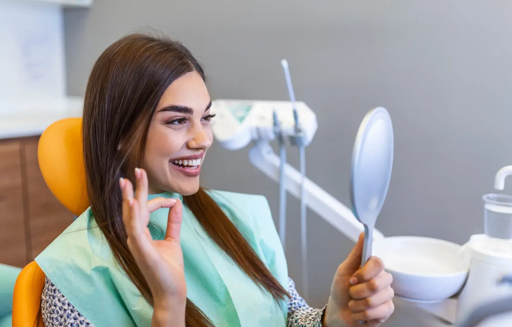 Smiling woman in dental chair holding a mirror and showing an OK gesture after dental treatment.
