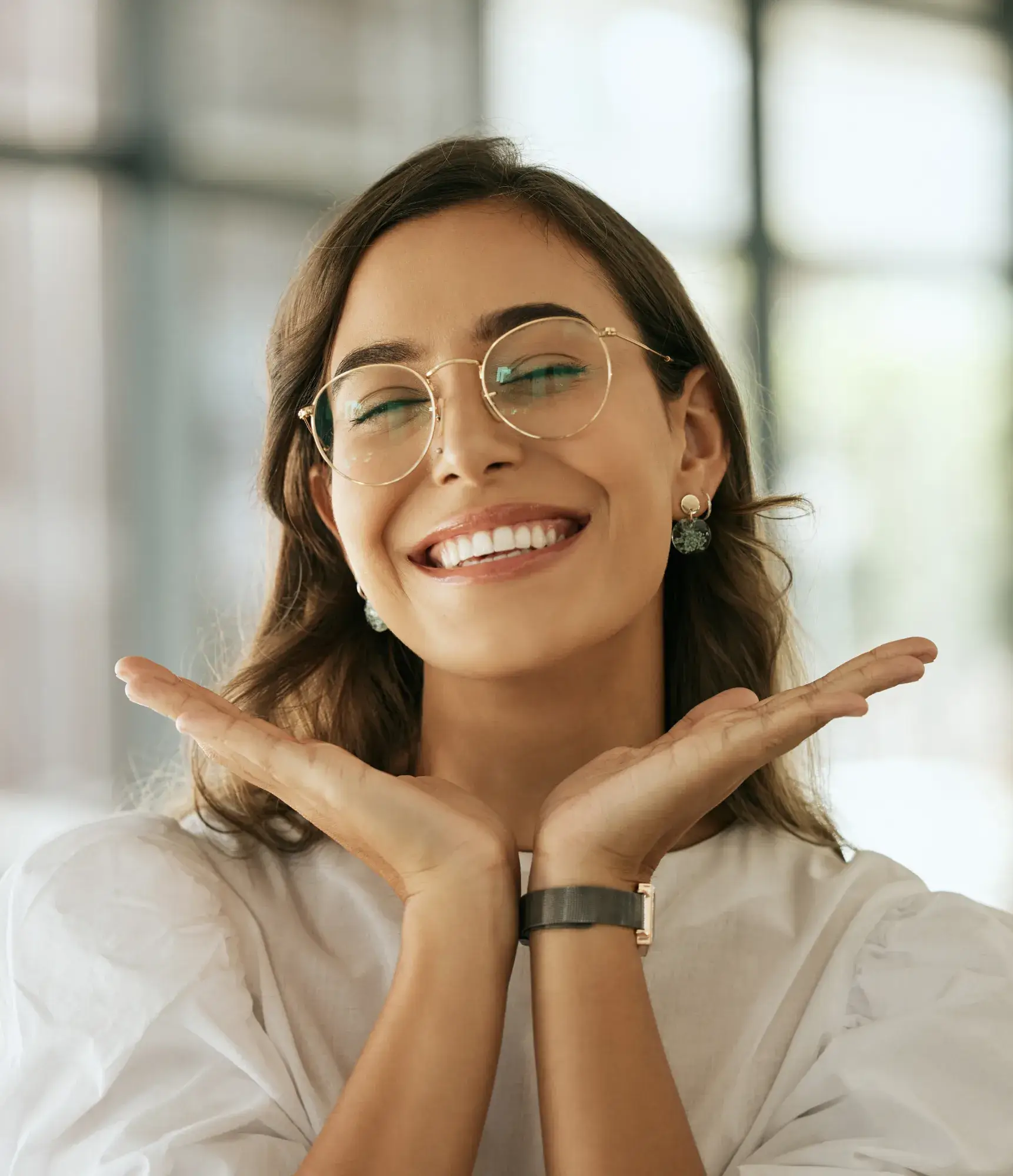 Smiling woman with glasses and earrings, hands framing her face in a bright indoor setting.