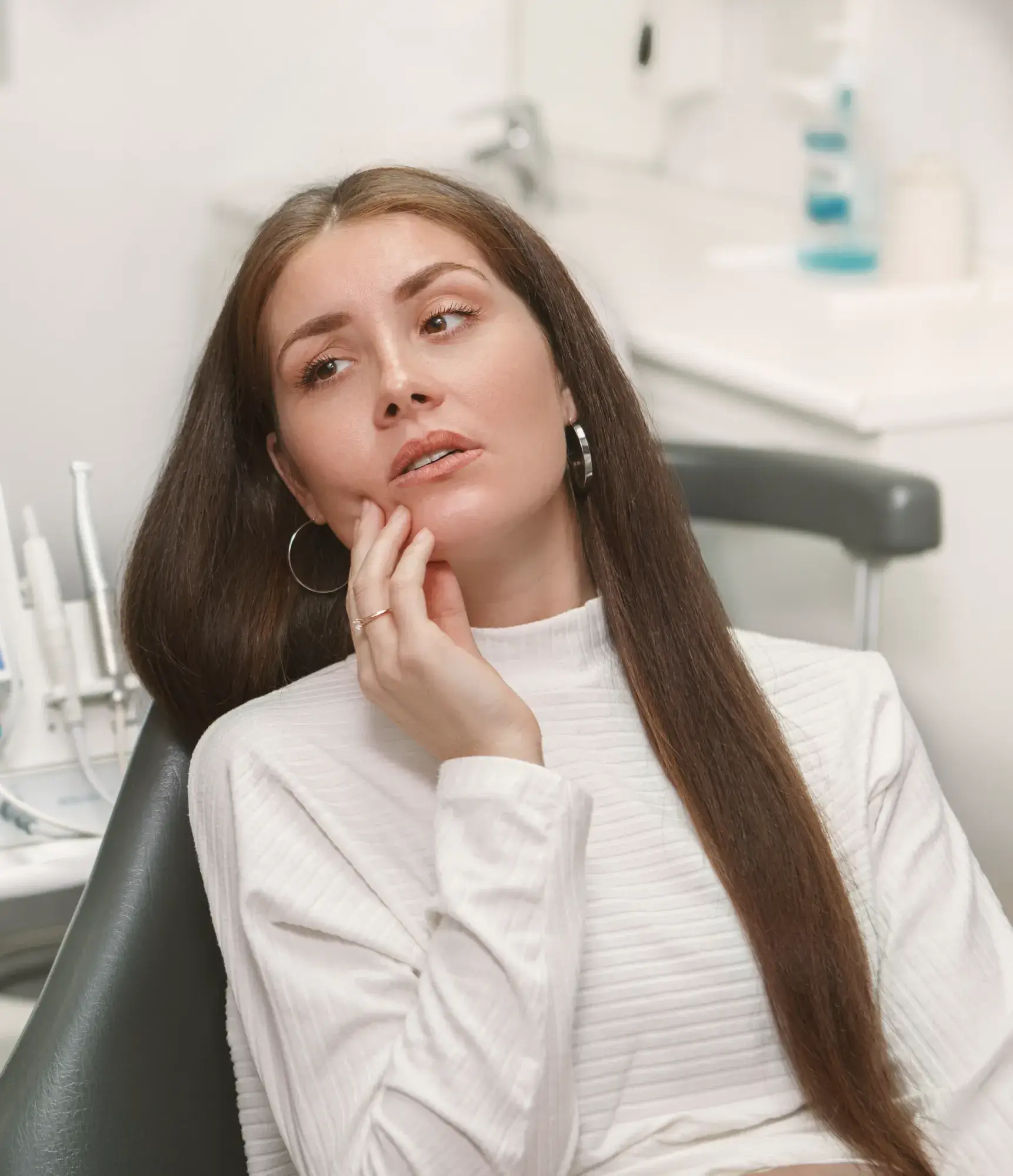 Woman with long brown hair sitting in a dental chair, holding her cheek with a pained expression.