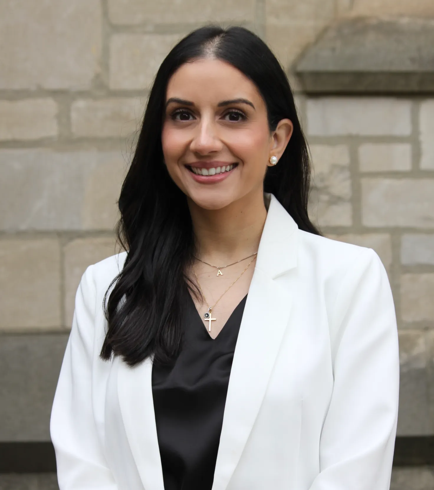 Smiling woman with long black hair wearing pearl earrings, two necklaces, a black top, and a white blazer standing in front of a stone wall.