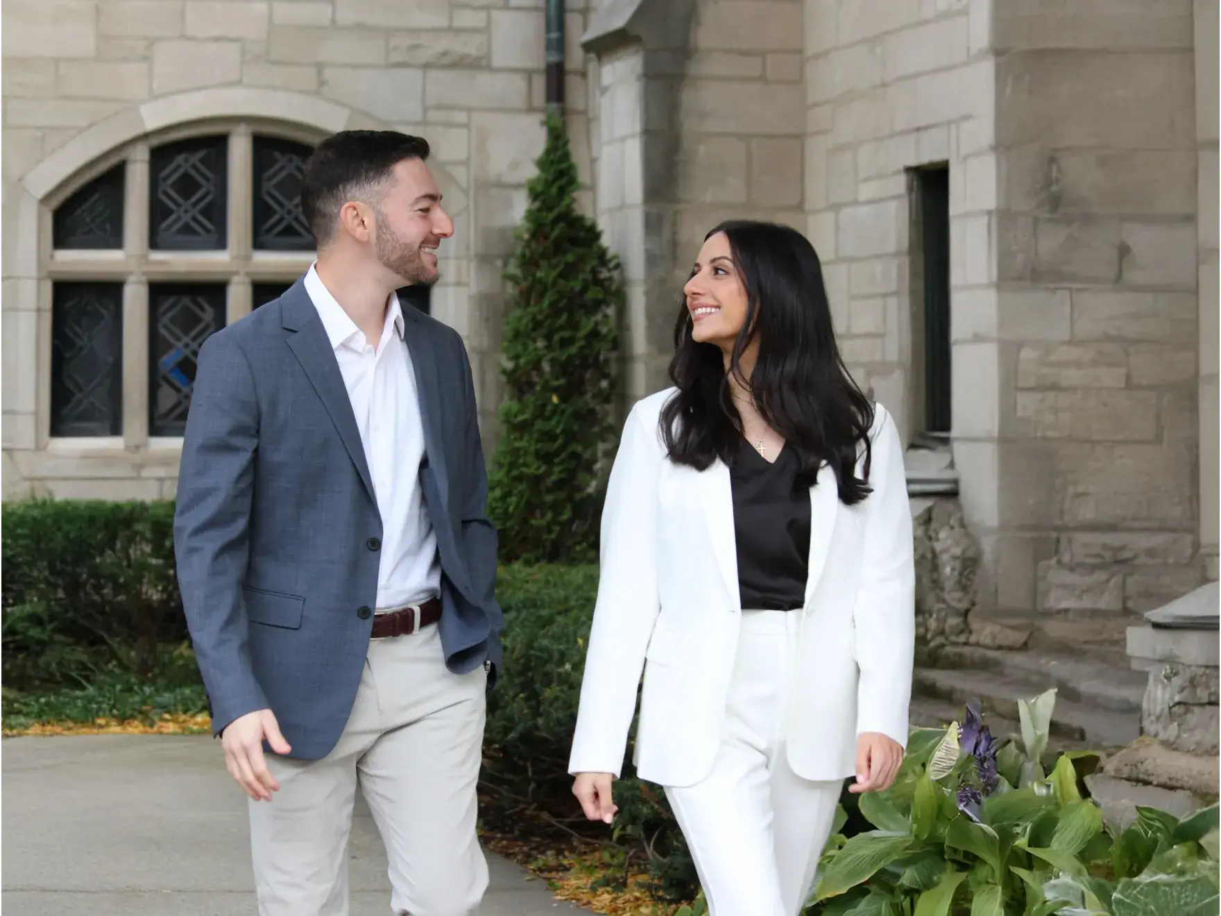 Man in gray blazer and beige pants smiling and walking while looking at woman in white suit walking beside him in front of stone building.