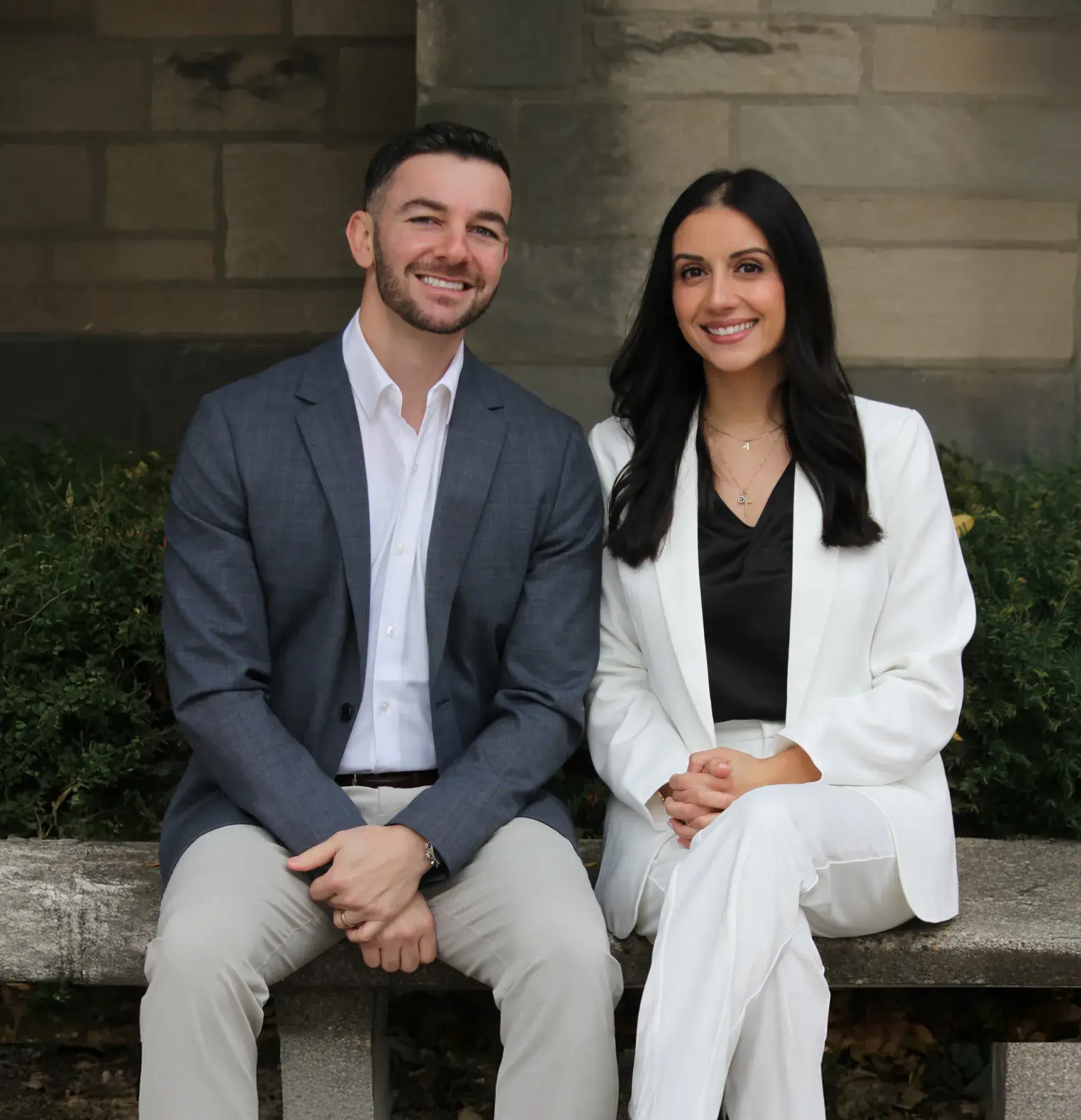 Smiling man in a gray blazer and beige pants sitting next to a woman in a white suit and black blouse on a stone bench outdoors.
