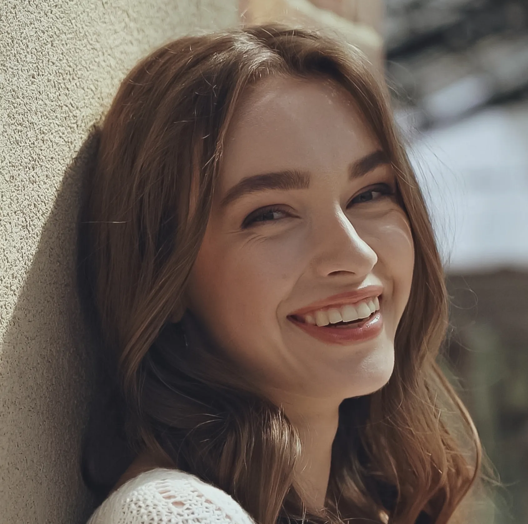 Young woman with long brown hair smiling and leaning against a textured wall outdoors.