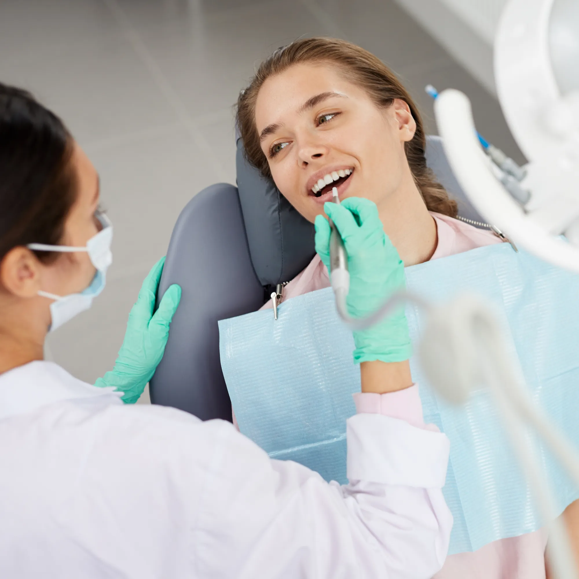 Dentist wearing green gloves and a mask examining a smiling patient’s teeth in a dental clinic.