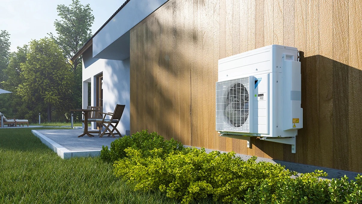 Outdoor air conditioning unit mounted on a wooden wall outside a modern house near shrubs and grass.