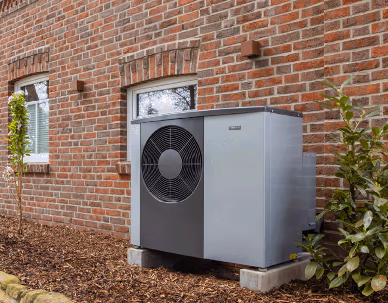 Outdoor heat pump unit mounted on concrete blocks next to a brick house with plants nearby.