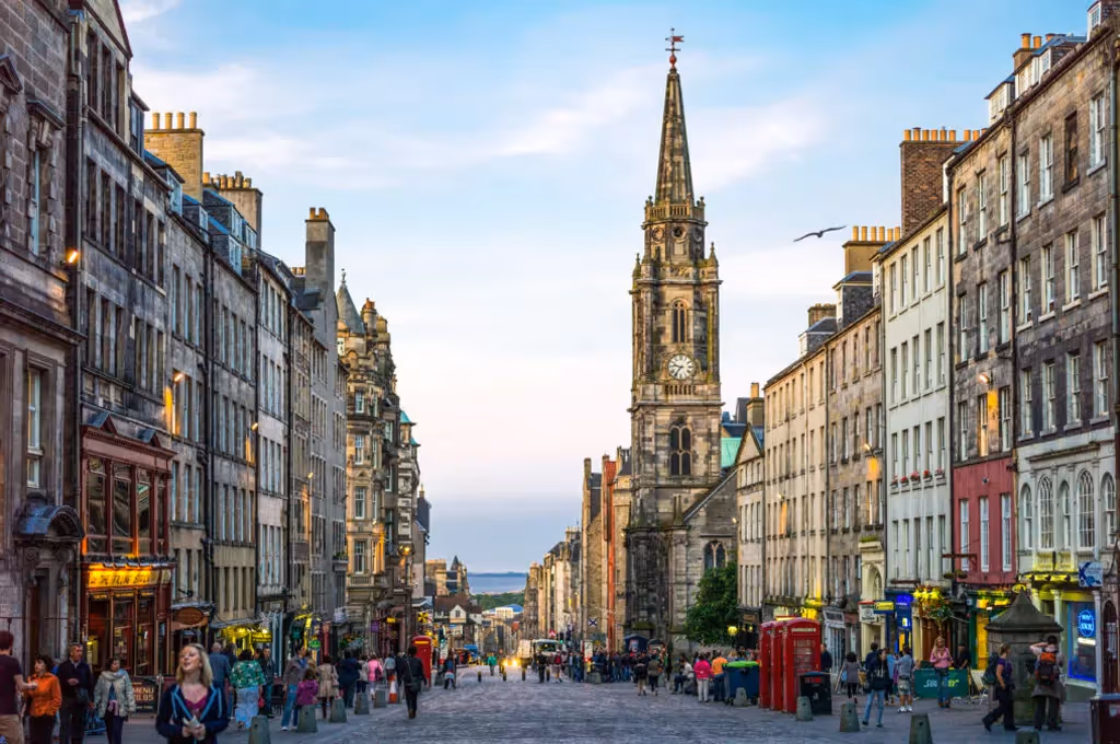 Crowded historic street with old stone buildings and a tall church steeple under blue sky.