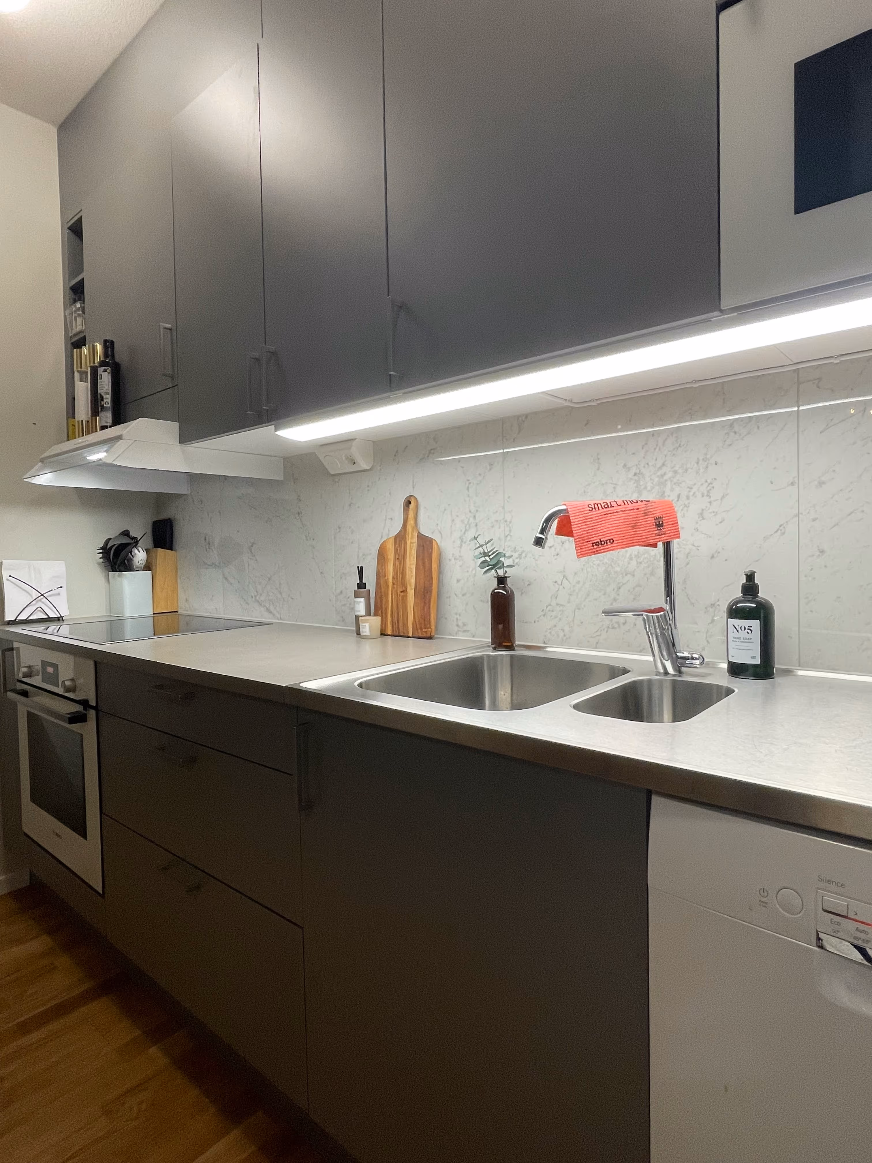 Modern kitchen with gray cabinets, stainless steel sink, oven, and under-cabinet lighting.