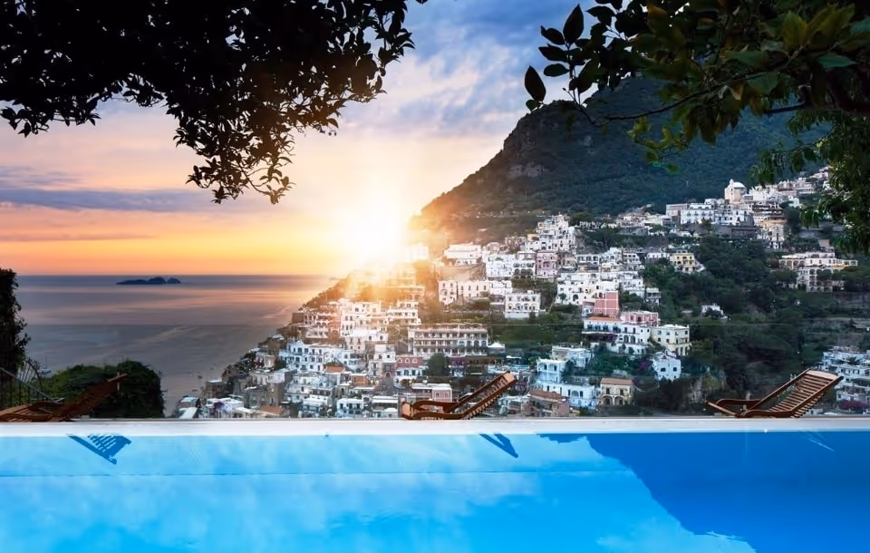 Sunset over a coastal town with white buildings on a hillside, viewed from a pool with lounge chairs and framed by tree branches.