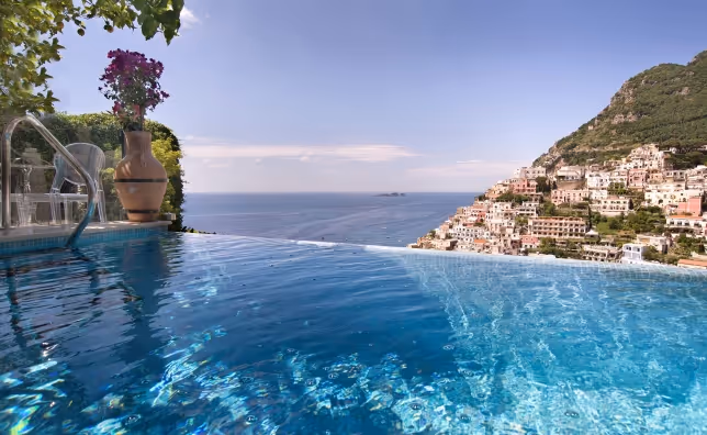 Infinity pool overlooking the sea with hillside town buildings in the background and a large terracotta pot with flowers on the poolside.