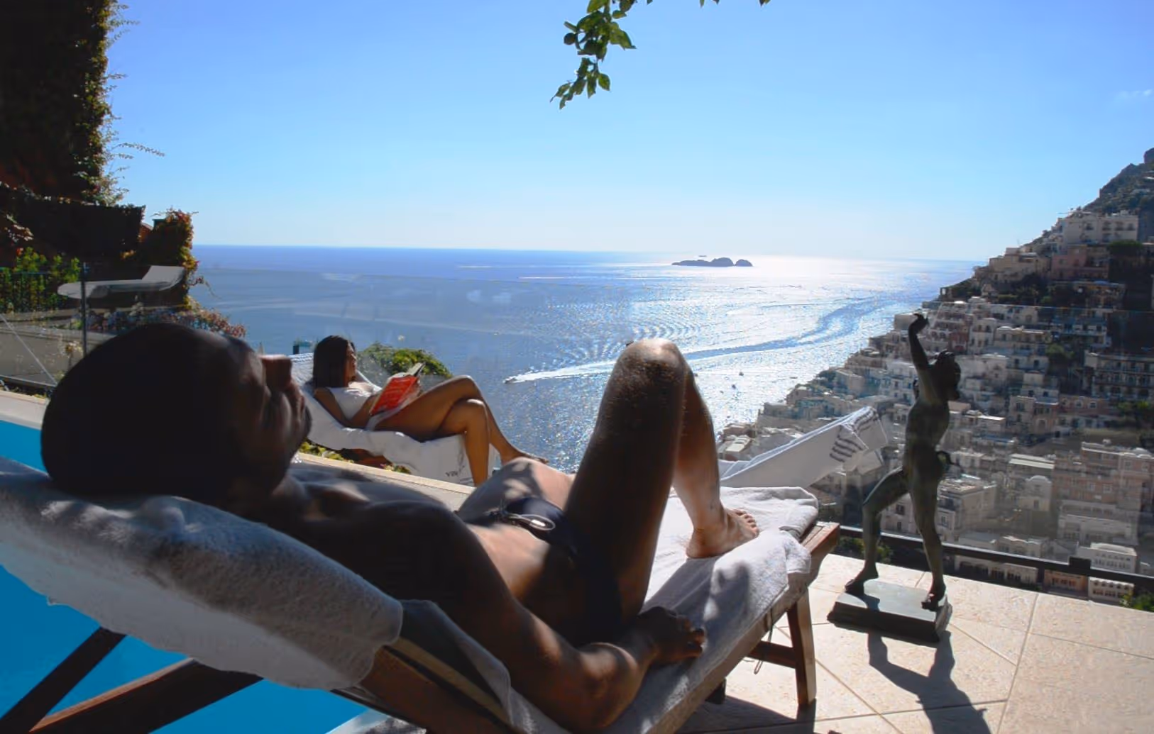 Two people relaxing on lounge chairs by a pool with a scenic view of the ocean and hillside town.