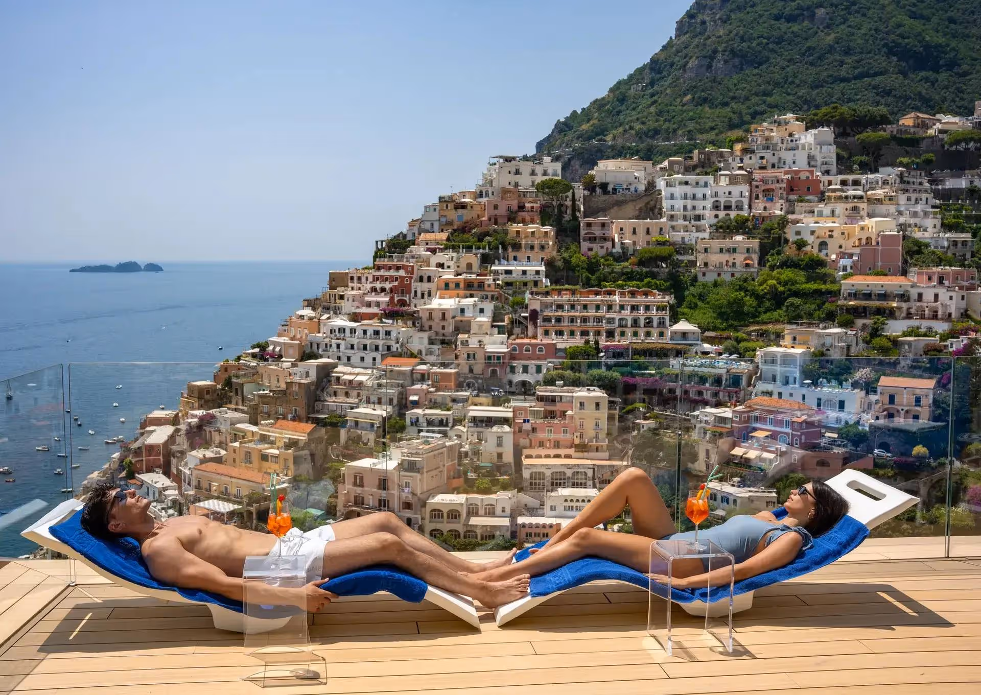 Man and woman relaxing on blue lounge chairs with orange drinks on a terrace overlooking colorful hillside buildings and the sea.