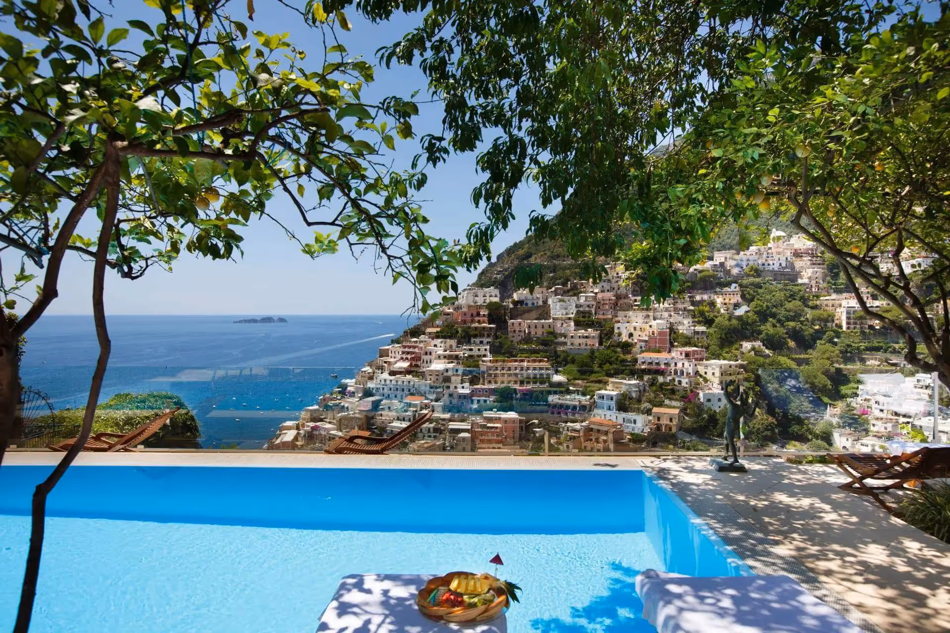 Infinity pool overlooking a coastal hillside town with colorful buildings, framed by green trees and a tray of fruit on a poolside table.