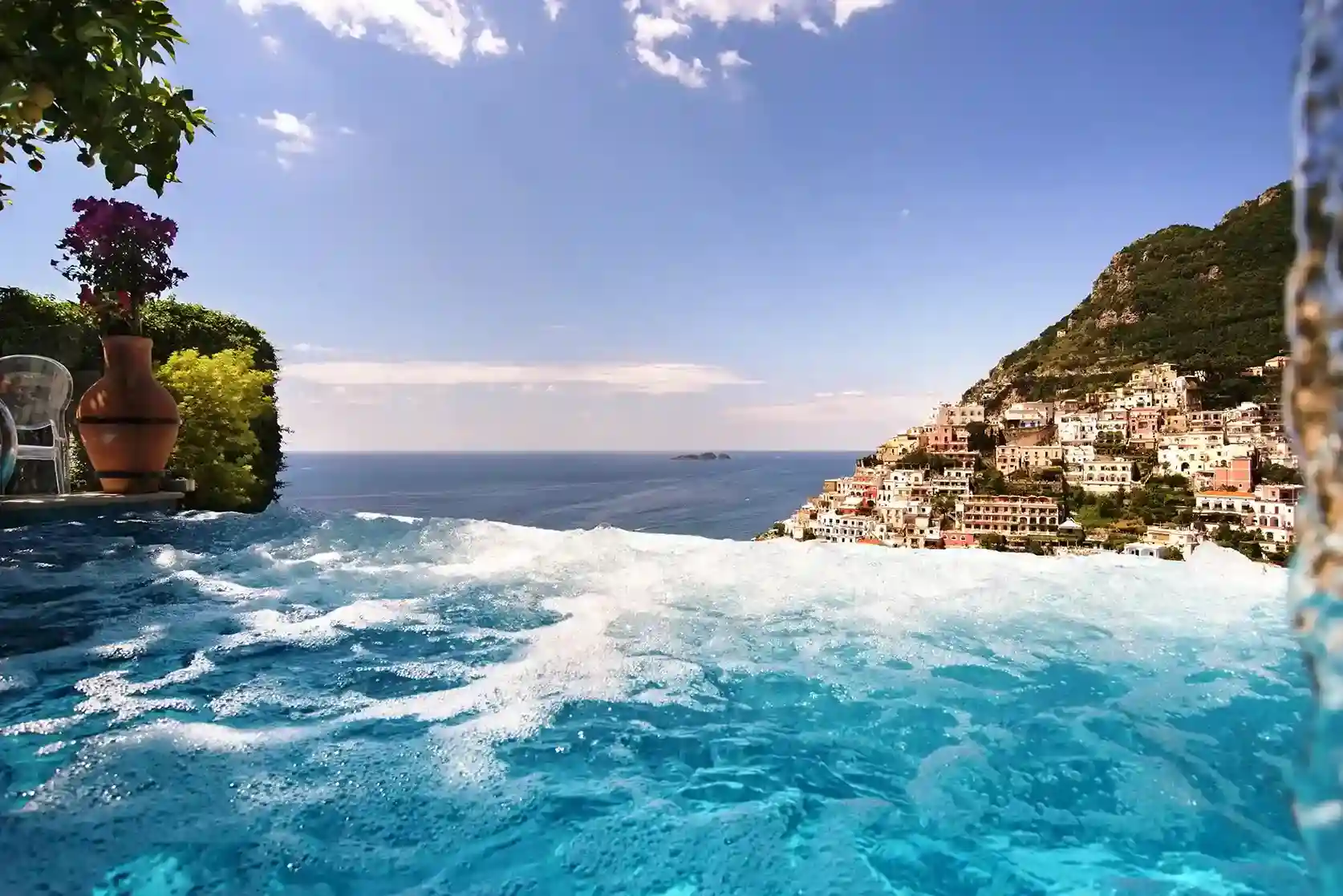 Bubbling infinity pool overlooking the sea and a hillside town under a blue sky.