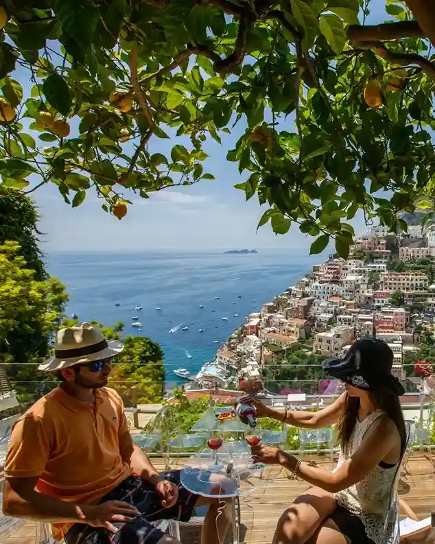Couple sitting on a terrace overlooking a coastal town, with lemons hanging overhead and the woman pouring red wine into glasses.