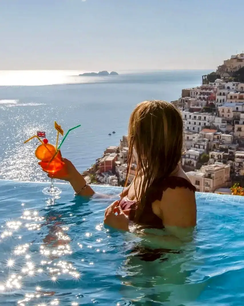 Woman with wet hair in an infinity pool holding a cocktail glass overlooking a coastal town and ocean at sunset.
