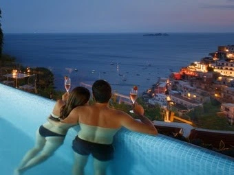 Couple in a pool raising glasses with a view of a coastal town and sea at dusk.