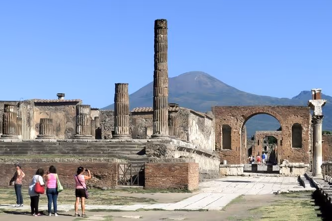 Tourists photographing ancient ruins with tall columns and an arch, with a mountain in the background under a clear blue sky.