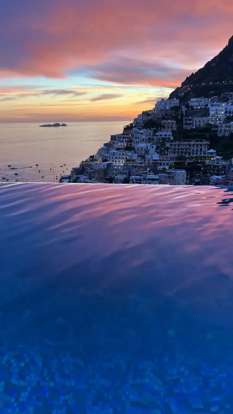 Infinity pool overlooking a coastal town at sunset with colorful sky reflections on the water.