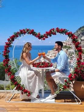 Couple sitting at a table, toasting with orange drinks under a heart-shaped rose arch overlooking a coastal town.
