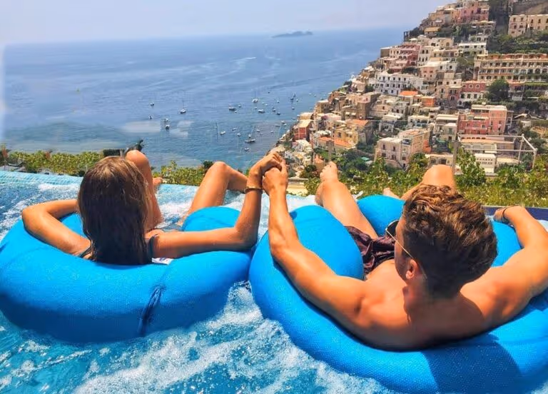 Couple floating on inflatables in the infinity pool, holding hands with Positano's colorful cliffside and the sea beyond