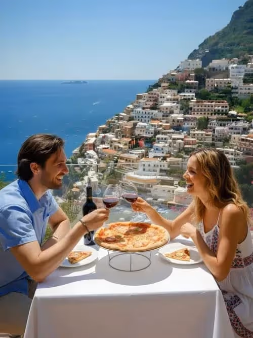 Couple clinking wine glasses over pizza overlooking a coastal town on the Amalfi Coast