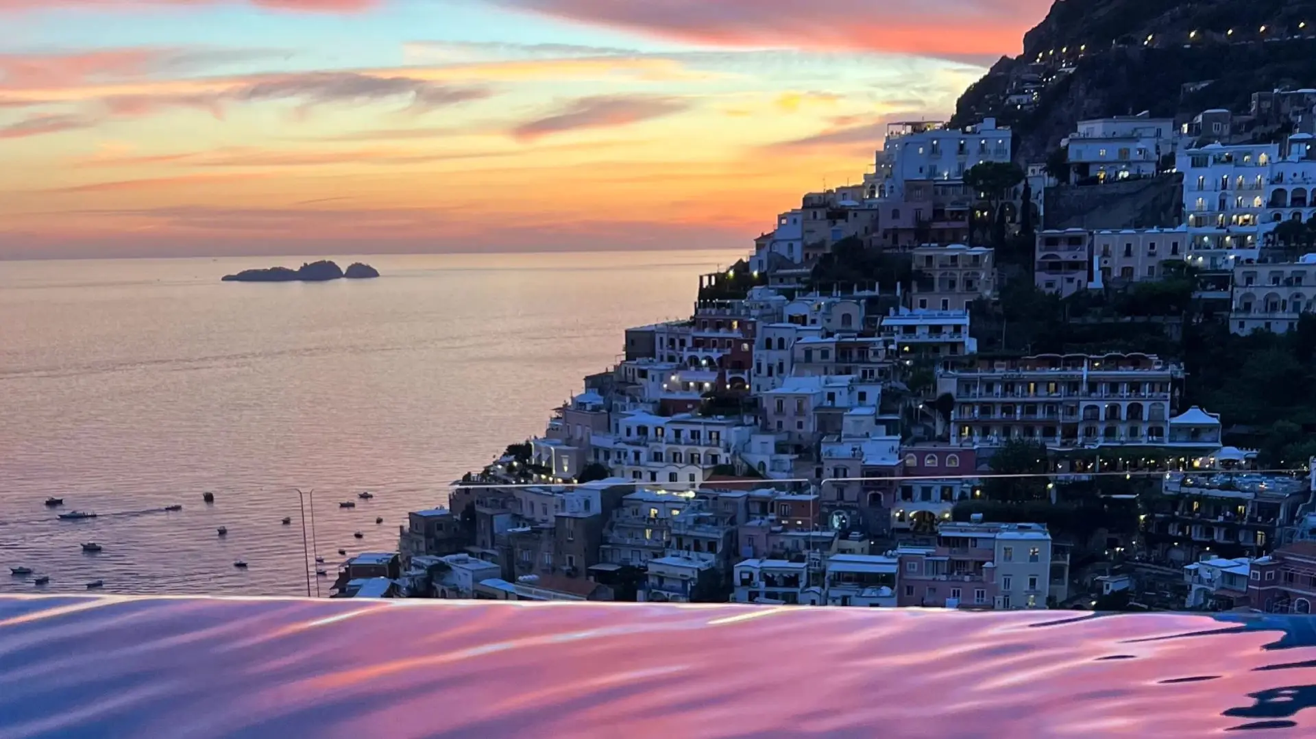 Sunset over Positano with boats on calm water reflected in an infinity pool