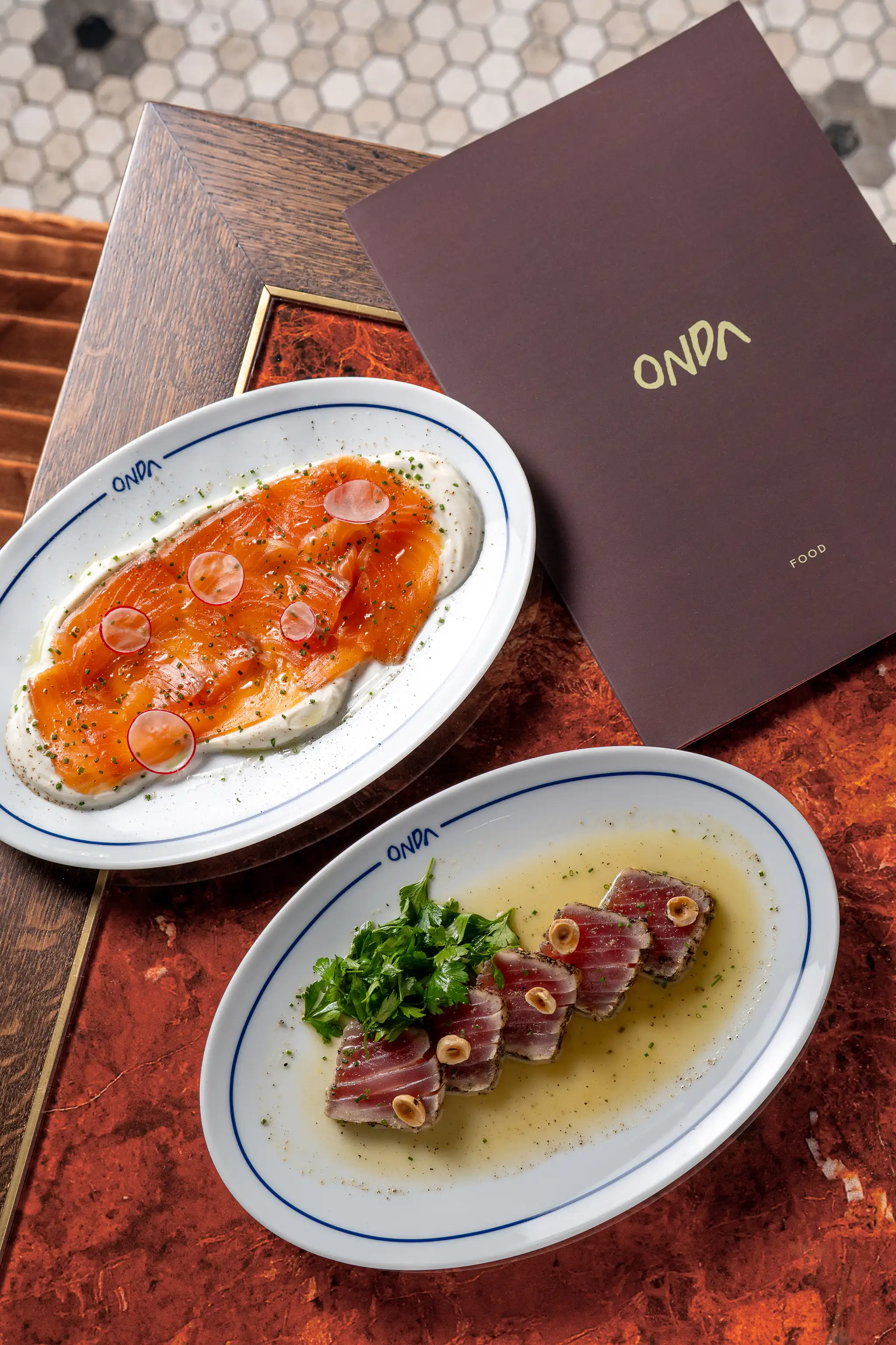 Two oval plates on a wooden and red marble table with a dark brown menu; one plate has thinly sliced smoked salmon with radish slices on a white creamy base, the other has seared tuna slices garnished with greens and nuts in a light sauce.